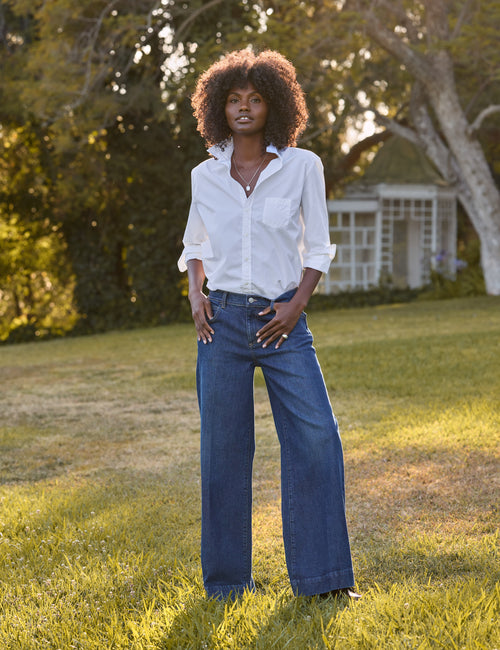 A woman stands outdoors on a grassy lawn wearing a white button-up shirt and dark blue wide-leg jeans, with trees and a small white structure in the background., view 4
