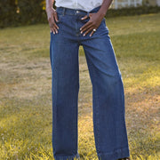 A woman stands outdoors on a grassy lawn wearing a white button-up shirt and dark blue wide-leg jeans, with trees and a small white structure in the background.