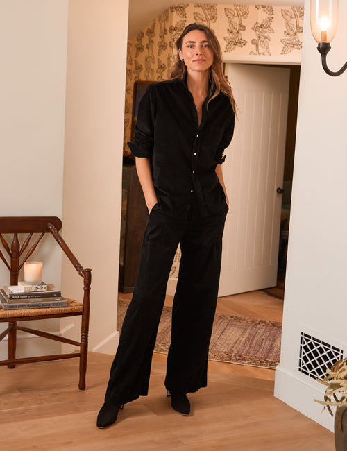 A woman stands in a warmly lit indoor room wearing a black long-sleeve button-front shirt and matching wide-leg pants, with her hands in her pockets., front view