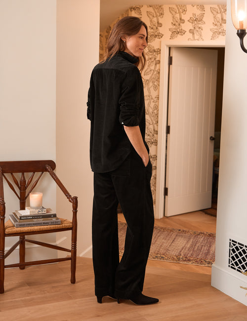 A woman stands in a warmly lit home interior wearing a black long-sleeve button-front shirt and matching black wide-leg trousers, shown from the back three-quarter view., alternate view