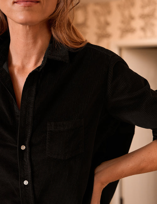 Close-up of a person wearing a black corduroy button-up shirt with a chest pocket, photographed indoors against a softly blurred neutral background., view 3