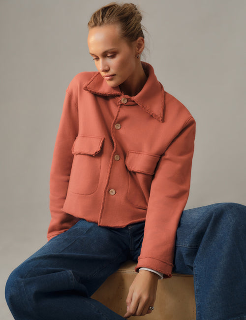 A woman wearing a terracotta-colored cropped jacket with a fuzzy collar and front buttons, paired with blue jeans, sitting on a wooden block against a neutral gray background., view 4