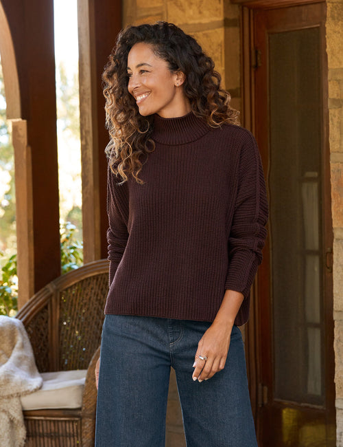 A woman stands on a stone porch wearing a dark brown ribbed turtleneck sweater, wide-leg blue jeans, and dark brown shoes., view 3