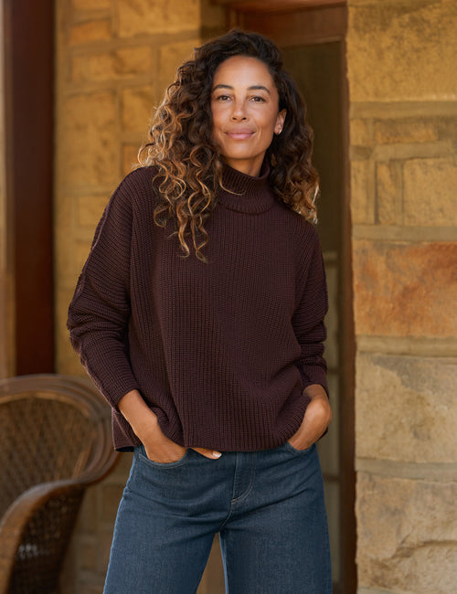 A woman with curly brown hair wearing a dark brown ribbed turtleneck sweater and blue jeans, standing in front of a stone wall., front view