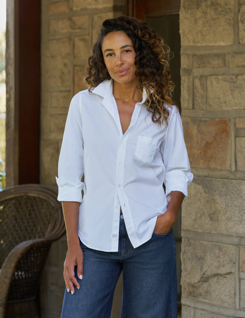 A woman with curly brown hair stands outdoors in front of a stone wall, wearing a white long-sleeve button-up shirt with rolled sleeves and dark blue jeans., front view