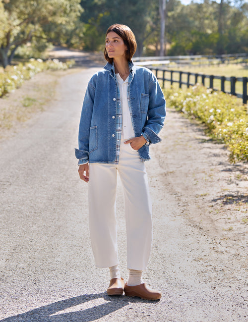 A woman stands on a sunlit gravel path outdoors wearing a light blue denim jacket over a white top and white wide-leg pants, with tan clogs and a watch., view 6