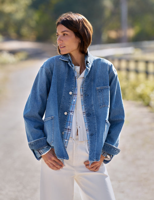 A woman wearing a light blue denim jacket over a blue plaid shirt and white pants, standing outdoors on a sunlit path with a blurred natural background., front view