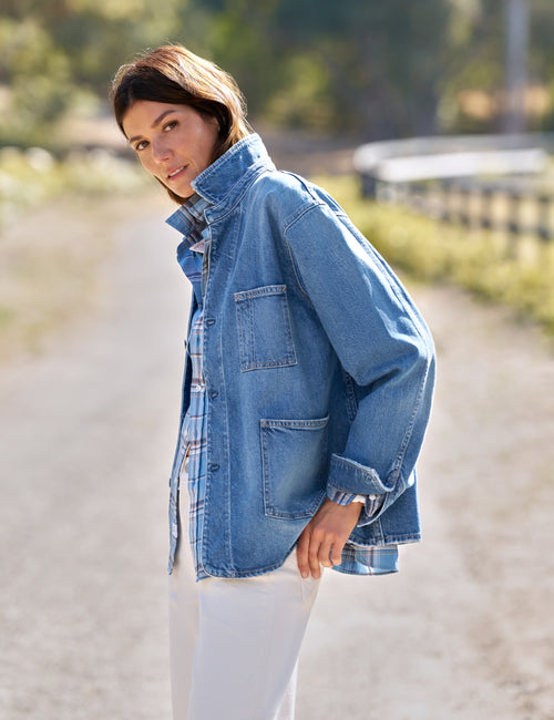 A woman wearing a light blue denim jacket over a plaid shirt and white pants, standing on a sunlit dirt path outdoors., alternate view