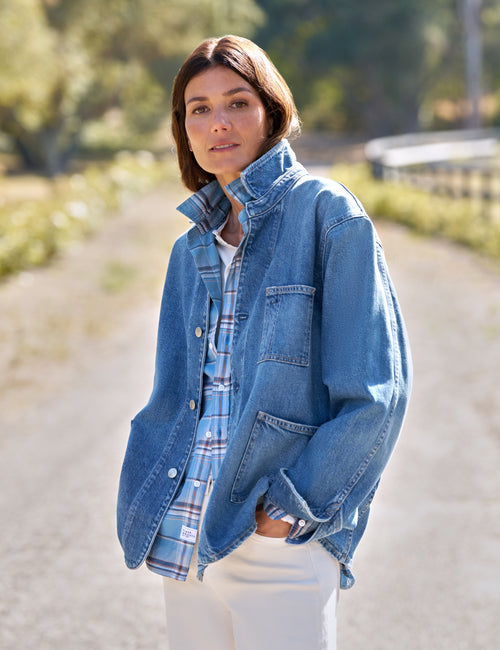 A woman wearing a light blue denim jacket over a blue and white plaid button-up shirt and white pants, standing on a sunlit dirt path outdoors., view 5