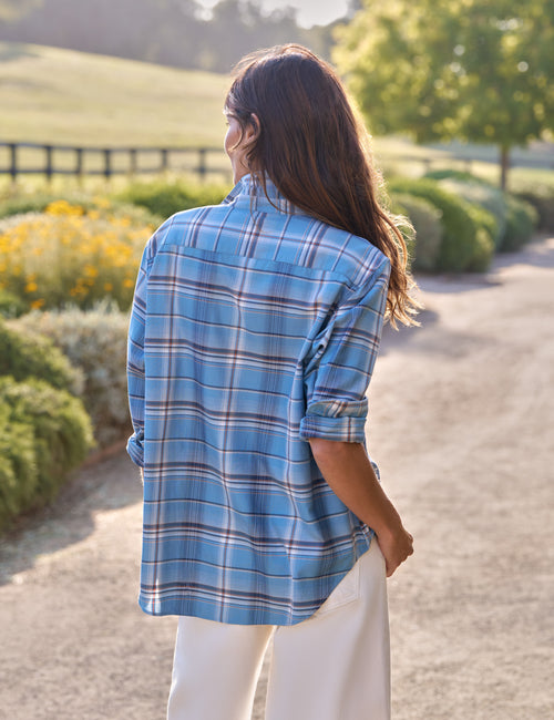 A woman viewed from behind wearing a light blue plaid button-up shirt with white pants, standing on a sunlit path in a park-like setting with greenery and flowers in the background., alternate view