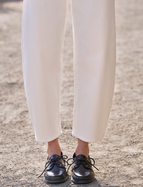 A close-up of a person's legs wearing off-white cropped trousers and shiny black lace-up shoes, standing on a sandy ground., view 3