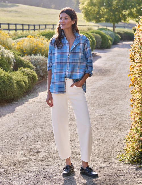 A woman stands on a sunlit gravel path outdoors, wearing a blue plaid button-up shirt, white cropped pants, and black shoes., view 4