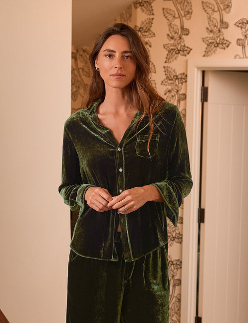 A woman stands indoors wearing a dark green velvet matching lounge set with a long-sleeve button-front top and wide-leg pants. The room has light wood flooring, patterned wallpaper, a chair with a candle, and a wall sconce., front view