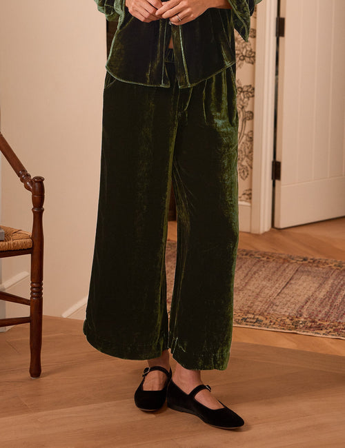 A woman stands indoors wearing a dark green velvet matching lounge set with a long-sleeve button-front top and wide-leg pants. The room has light wood flooring, patterned wallpaper, a chair with a candle, and a wall sconce., front view