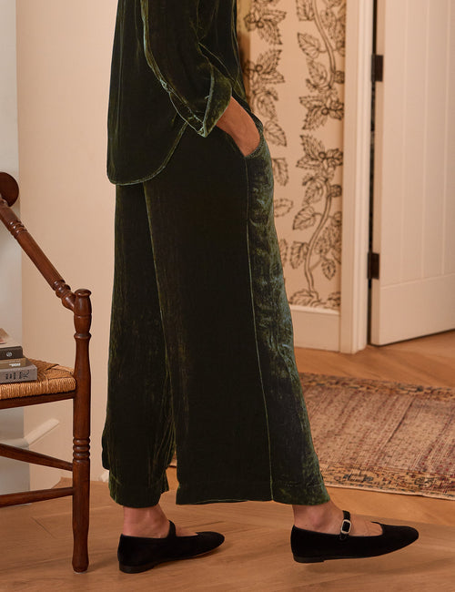 A woman wearing a dark green velvet two-piece set with a high-neck long-sleeve top and matching wide-leg trousers, standing in a warmly lit room with patterned wallpaper, a wooden chair, and a lit candle., alternate view