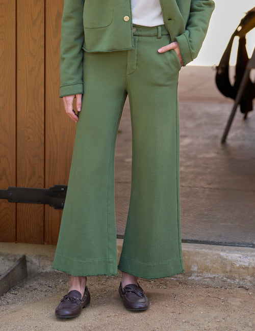 A person wearing an olive green tailored pantsuit with a cropped jacket and wide-leg trousers, paired with a white top and dark brown loafers, standing outdoors against a wooden door and concrete ground., front view
