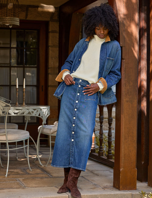 A woman stands on a porch wearing a blue denim jacket with tan collar and cuffs, a cream turtleneck, a long blue denim button-front skirt, and brown boots., view 5