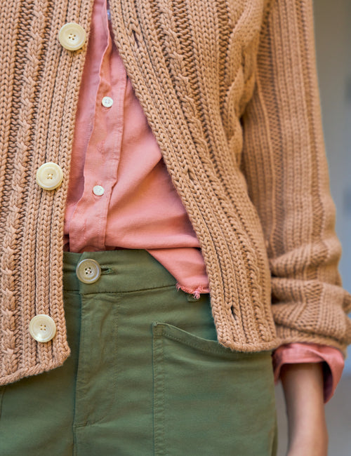 A close-up of a person wearing a chunky knit cardigan in a warm tan/beige color over a pink button-up shirt and olive green pants., alternate view