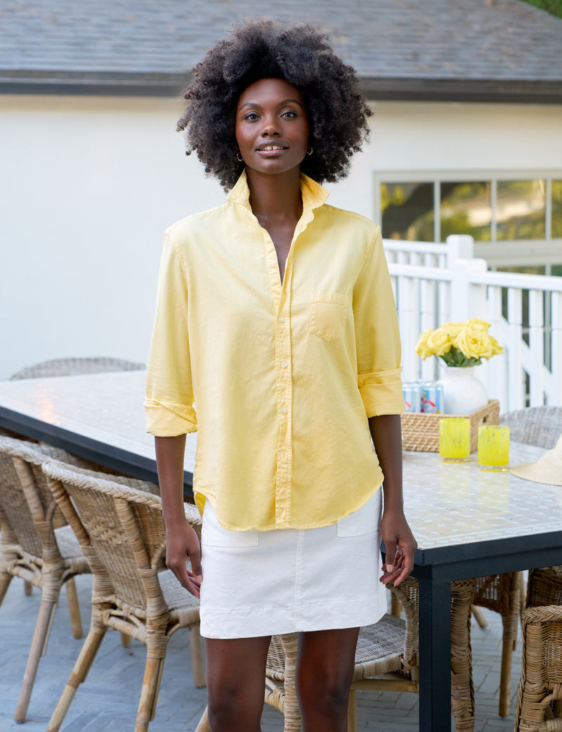Woman in a yellow shirt and white skirt standing outdoors near a dining table with chairs.