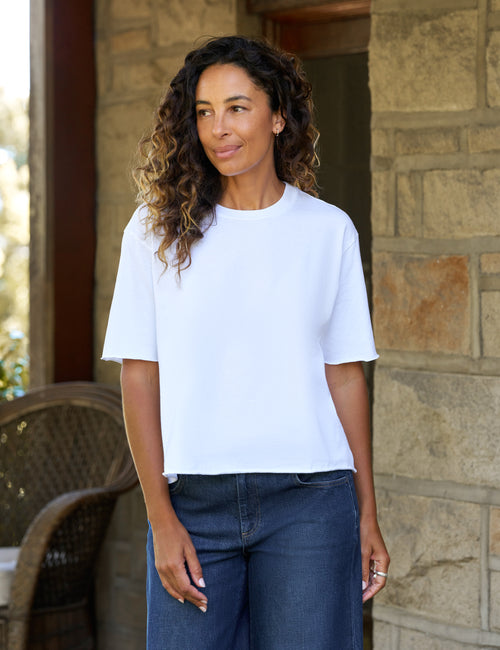 A woman with curly brown hair wearing a white short-sleeve crewneck t-shirt and dark blue jeans, standing in front of a stone wall., front view
