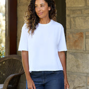 A woman with curly brown hair wearing a white short-sleeve crewneck t-shirt and dark blue jeans, standing in front of a stone wall.