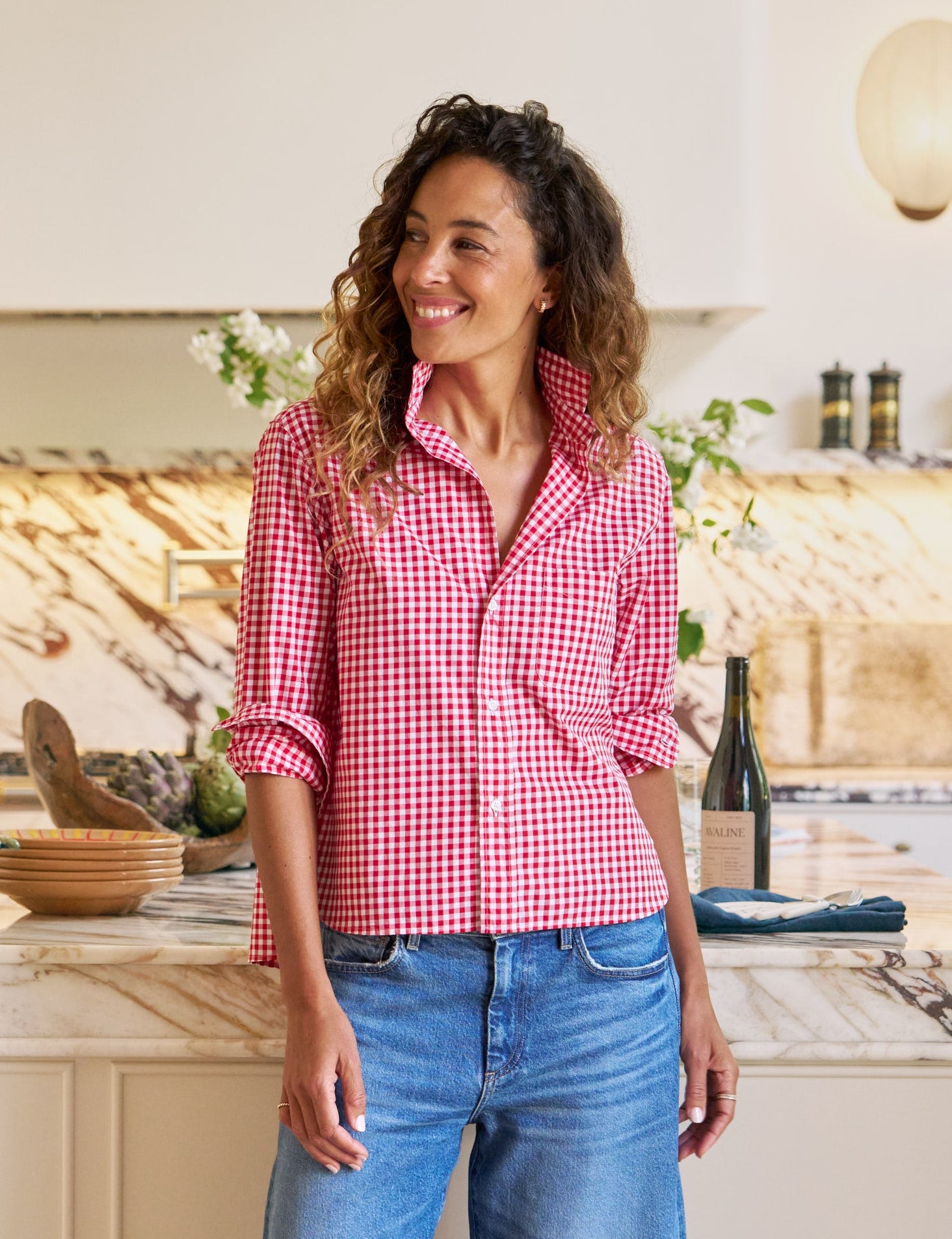 Woman standing in a kitchen wearing a red checkered shirt and blue jeans.