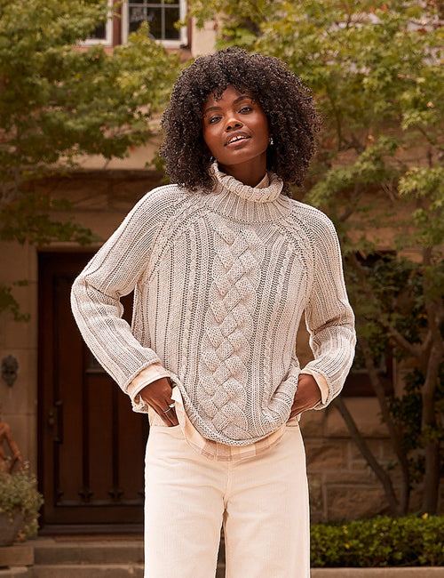 A woman stands outdoors in front of a stone house wearing a cream cable-knit turtleneck sweater, cream wide-leg pants, and dark brown leather shoes., front view