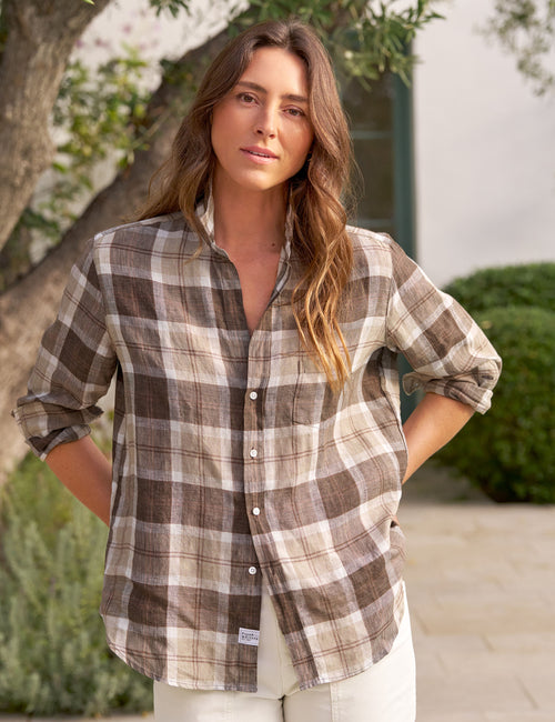 A woman wearing a brown and beige plaid button-down shirt with rolled-up sleeves, standing outdoors in front of a tree and stone wall., front view