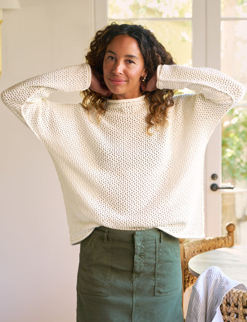 A woman wearing a cream-colored, long-sleeve textured knit sweater and an olive green mini skirt, standing in a bright, modern room with a window and wicker furniture in the background., view 4