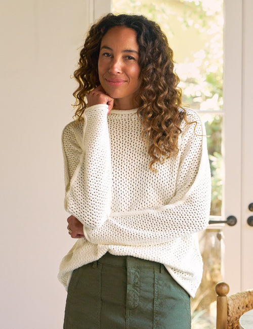 A woman with curly brown hair stands indoors wearing a cream open-knit long-sleeve sweater and an olive green skirt. She is posed in front of white French doors with a woven chair and table visible in the softly lit room., front view