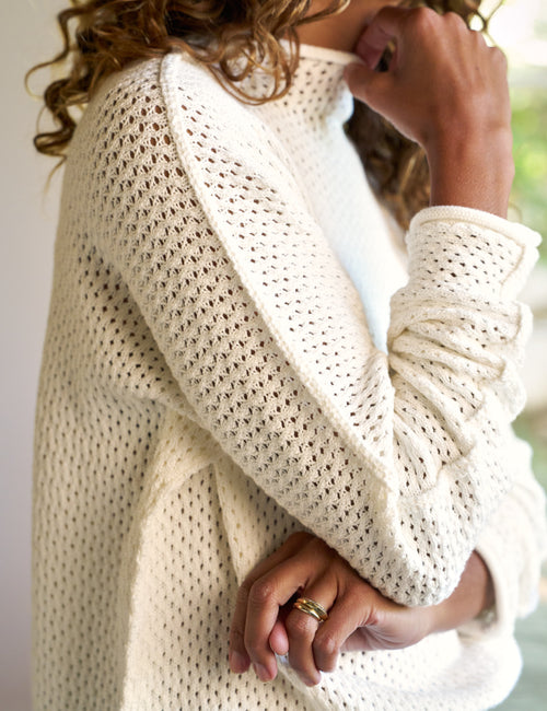 A close-up of a person wearing a cream-colored, open-knit long-sleeve sweater. The image highlights the textured knit pattern and the soft drape of the fabric, with the person's hands and hair visible against a softly blurred light background., alternate view