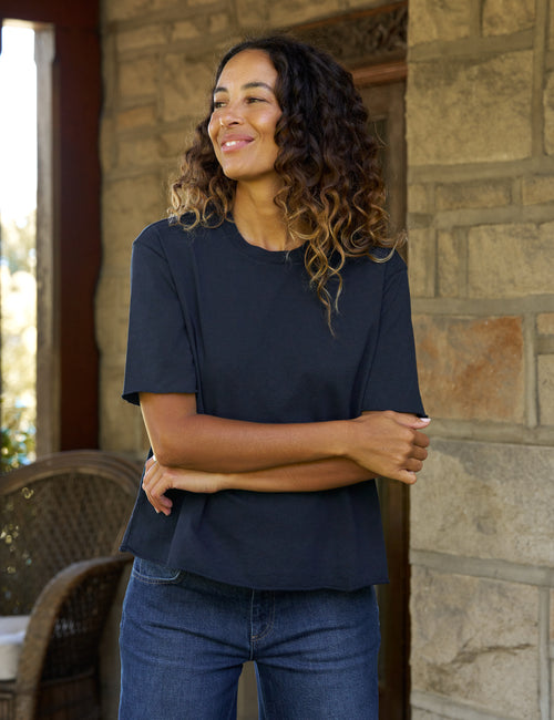 A woman with curly brown hair wearing a dark navy blue short-sleeve t-shirt and blue jeans, standing on a stone porch with her arms crossed., front view