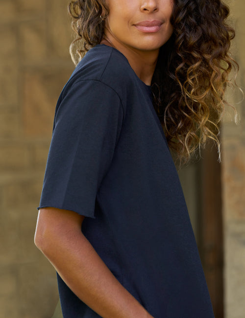 A woman wearing a dark navy blue short-sleeve t-shirt, shown from a side-back angle against a blurred stone wall background., view 3