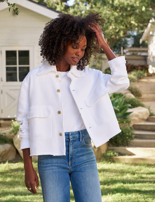 A woman wearing a white cropped jacket with gold buttons and blue jeans, standing outdoors in a garden setting., view 3
