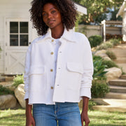 A woman wearing a white cropped jacket with front buttons and flap pockets, paired with blue jeans, standing outdoors in a garden setting.