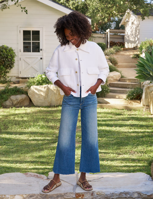 A woman stands outdoors in a grassy yard wearing a white cropped button-front jacket with front flap pockets, blue wide-leg cropped jeans, and brown sandals., view 5