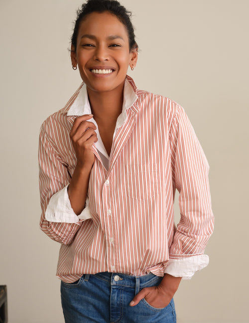 A smiling woman wearing a orange and white vertically striped button-down shirt with white rolled cuffs, paired with blue jeans, against a plain beige background., view 5