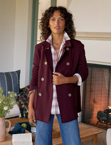 Woman wearing a burgundy coat with a plaid shirt and blue jeans in a cozy living room.