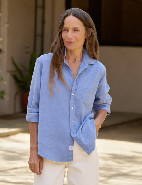 A woman wearing a light blue long-sleeve button-up shirt with rolled-up sleeves and white pants, standing outdoors in front of a neutral-toned building., front view