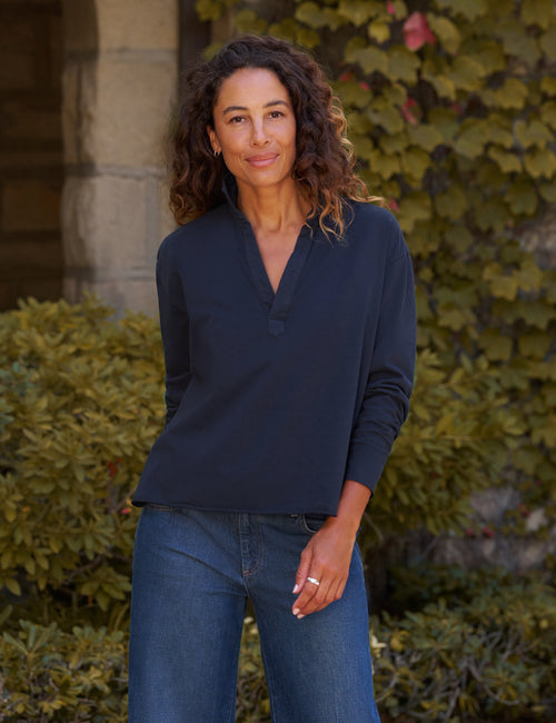 A woman with curly brown hair wearing a dark navy long-sleeve henley shirt and blue jeans, standing outdoors in front of a stone column and green ivy-covered wall., front view