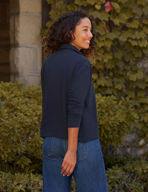 A woman with curly dark hair is shown from the back and side, wearing a dark navy long-sleeve button-up shirt and blue jeans, standing outdoors in front of green ivy-covered walls., alternate view