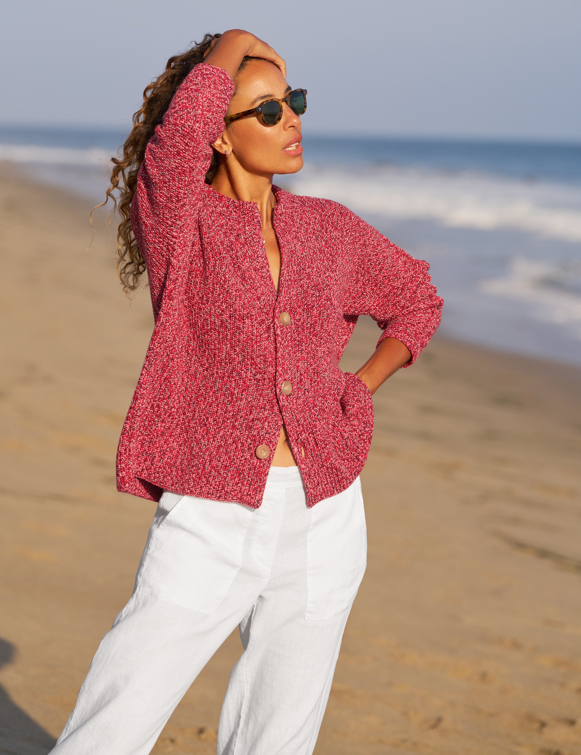 Woman in a red knitted cardigan and white pants standing on a beach., alternate view