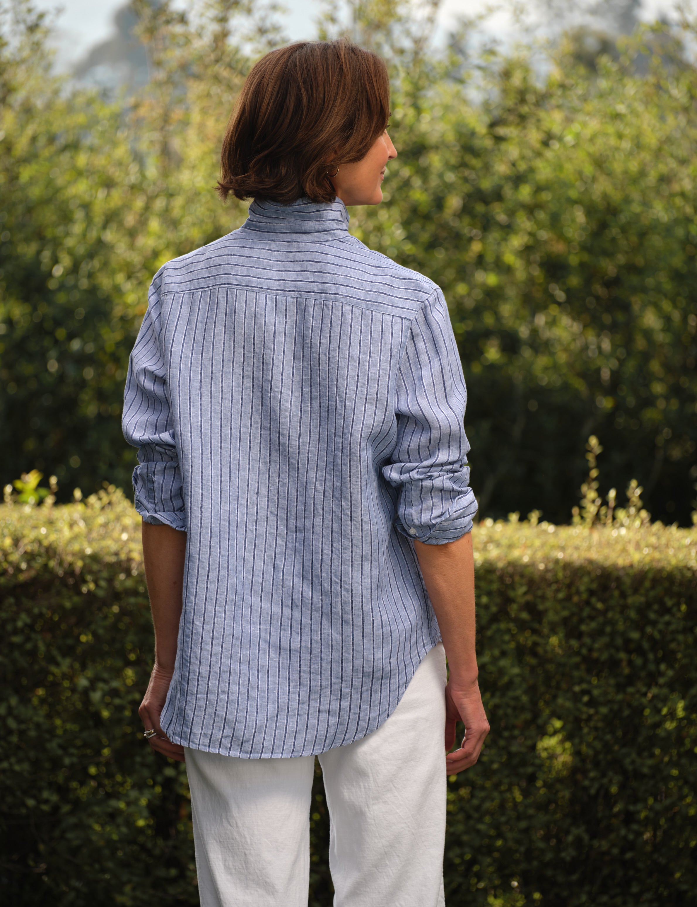 Person wearing a blue striped shirt and white pants standing outdoors with greenery in the background, alternate view