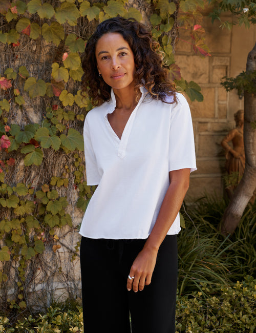 A woman wearing a white short-sleeve V-neck blouse and black pants, standing outdoors in front of a stone wall covered in green and yellow vines., front view