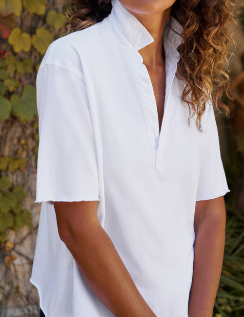 A woman wearing a white short-sleeve collared blouse with a split placket, photographed outdoors against a blurred background of foliage and tree bark., view 3