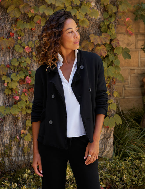 A woman wearing a black double-breasted jacket over a white collared shirt and black pants, standing outdoors in front of a stone wall covered in green and red autumn ivy., front view
