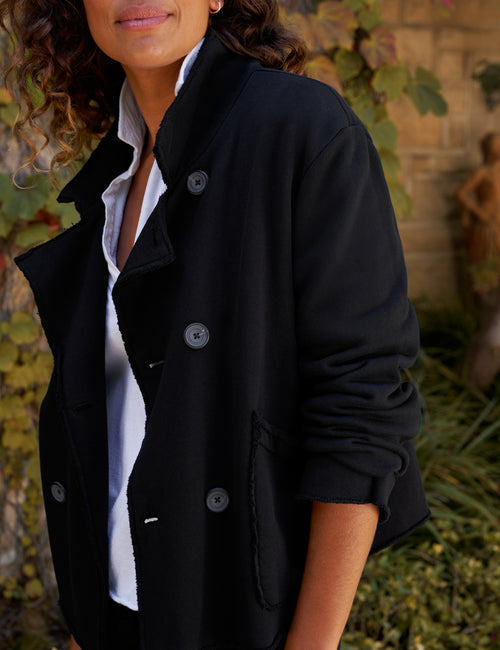 A woman wearing a black double-breasted coat over a white collared shirt, photographed outdoors with foliage and a stone wall in the background., view 3