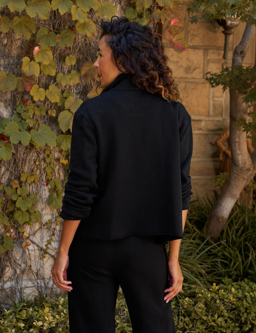 A woman with curly dark hair is shown from behind wearing a black cropped long-sleeve jacket and black pants, standing outdoors in front of a stone wall covered in green and yellow ivy., alternate view