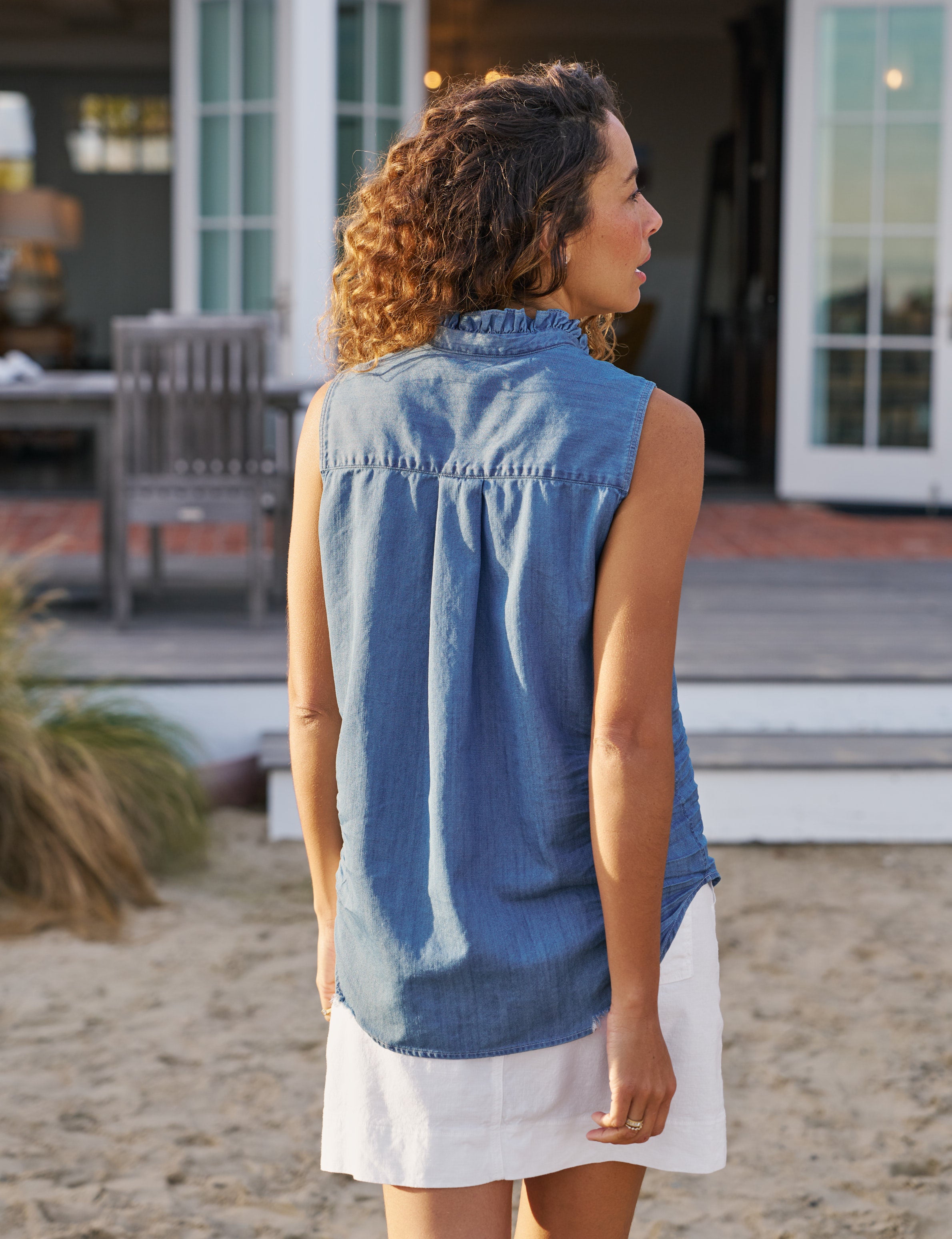 Woman wearing a blue sleeveless shirt and white skirt standing on a beach., alternate view
