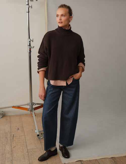 A model stands in a studio wearing a dark brown ribbed turtleneck sweater, dark blue barrel-leg jeans, and black loafers. A studio light stand is visible on the left against a neutral light gray backdrop., view 4